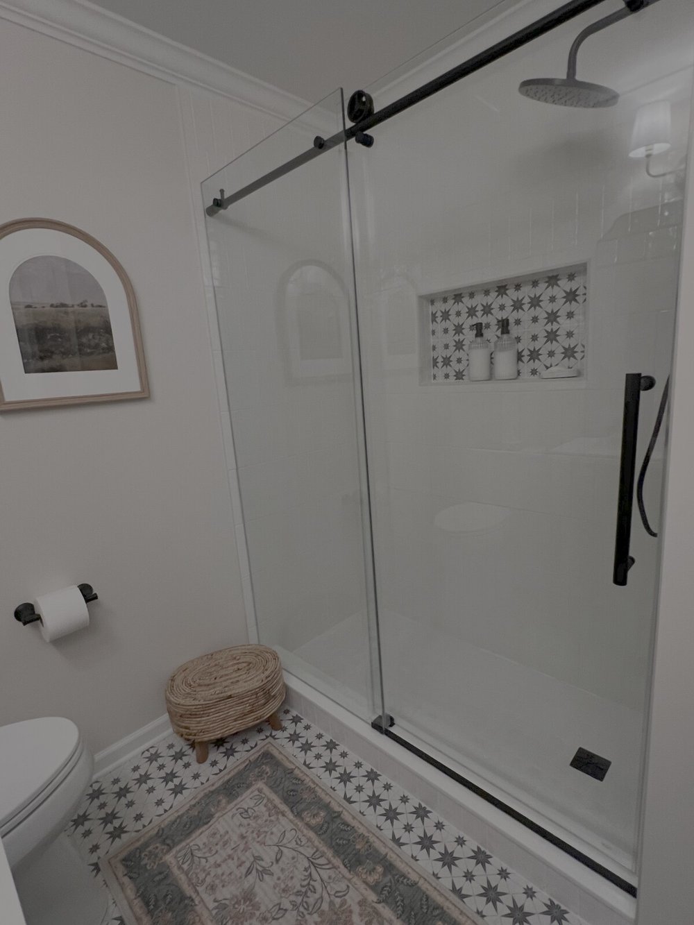 Completed white tiled shower with frameless sliding glass door, black hardware, patterned niche and the homeowner’s artwork and woven basket