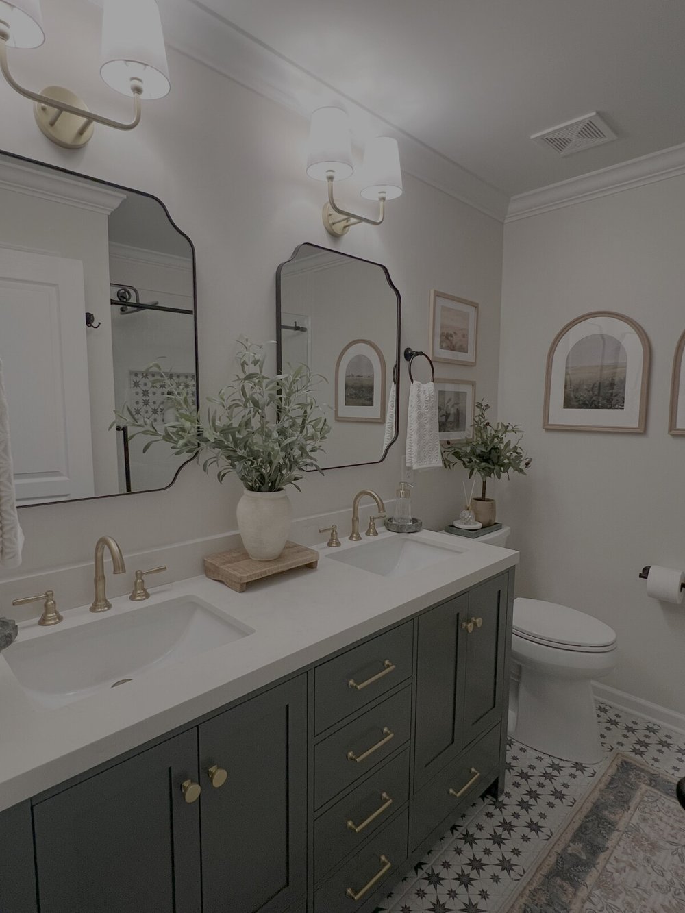 Green double vanity with brass faucets, shaped mirrors, wall sconces, greenery and the homeowner’s artwork and towels